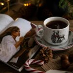 A cup of holiday-flavored tea sits next to an open magazine, a candy cane, and other holiday scenery.